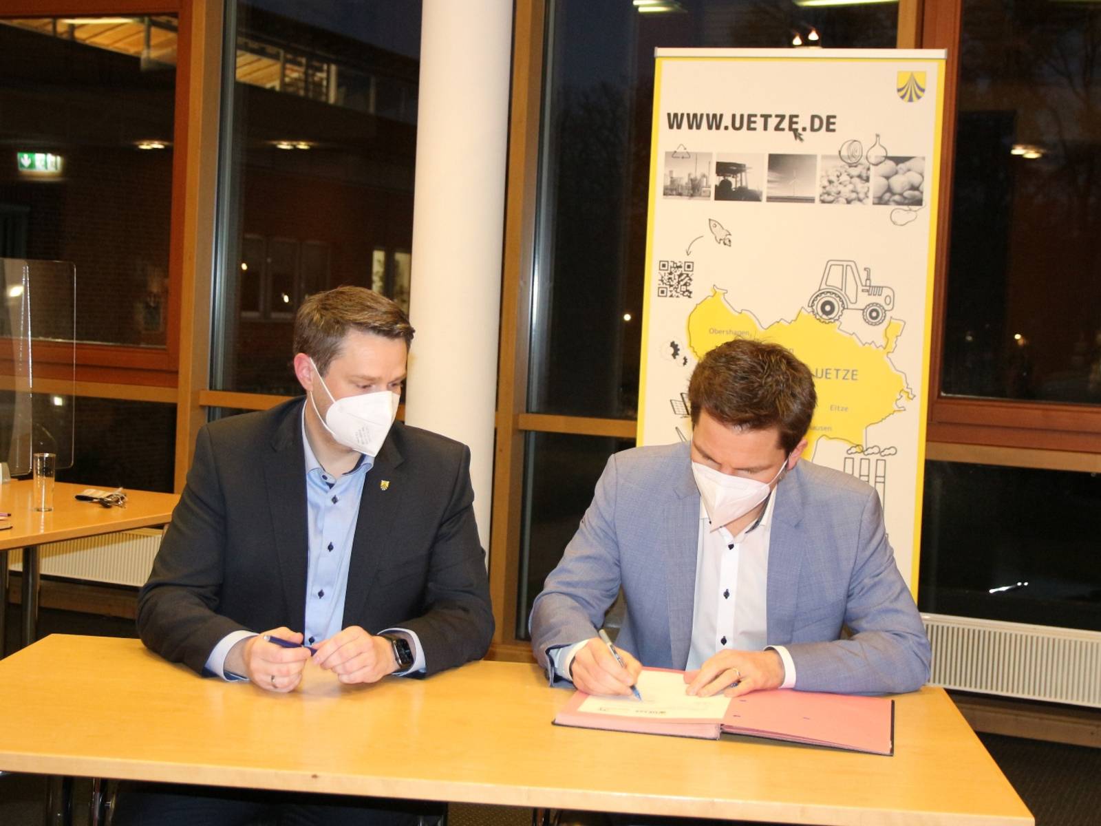 Zwei Herren an einem Tisch, einer unterschreibt  etwas. Der andere schaut zu. Im Hintergrund ein Banner mit der Aufschrift www.uetze.de.